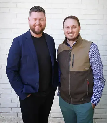 Stefan Bunch stands beside Drew Sanford, smiling in front of a white brick wall.